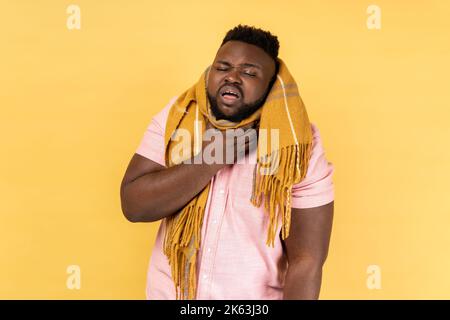 Ritratto dell'uomo che indossa una camicia rosa in una sciarpa calda che si sente malata con mal di gola, febbre alta temperatura, sintomi influenzali stagionali. Studio al coperto isolato su sfondo giallo. Foto Stock