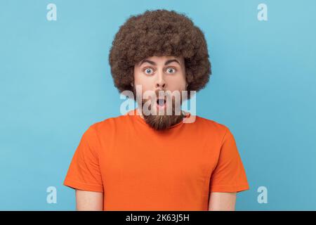 Ritratto di uomo stupito con acconciatura afro indossare T-shirt arancione in piedi con la bocca aperta di sorpresa, ha scioccato l'espressione, sente incredibili notizie. Studio in interni isolato su sfondo blu. Foto Stock