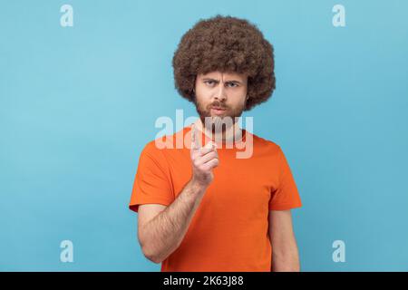 Ritratto di uomo serio con acconciatura afro indossare T-shirt arancione in piedi con gesto admonishing, scolding per errore e avvertimento. Studio in interni isolato su sfondo blu. Foto Stock