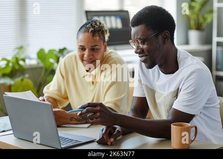 Immagine laterale di due persone nere sorridenti che utilizzano il notebook insieme in ufficio IT e indossano abiti informali che puntano sullo schermo Foto Stock