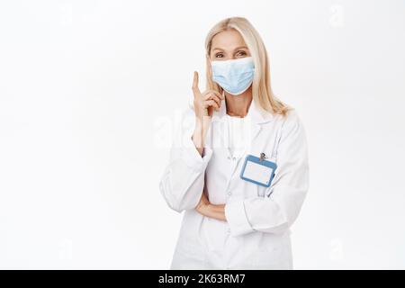 Ritratto di un operatore sanitario sorridente, dipendente di laboratorio in maschera medica, punta il dito verso l'alto, mostra la pubblicità, si trova su sfondo bianco Foto Stock