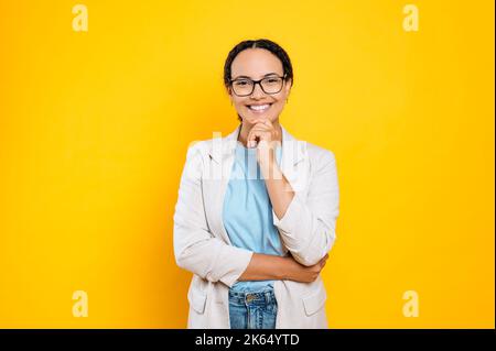 Bella bella positiva giovane brunetta brasiliana o donna ispanica con occhiali, vestiti elegantemente, dipendente aziendale, in posa in piedi su sfondo isolato arancione, guarda la macchina fotografica, sorridendo Foto Stock