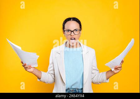 Confuso perplesso preoccupato giovane latino o donna brasiliana, donna d'affari, capo, tenere documenti in mano, tenere la bocca aperta, guardare la macchina fotografica irritata, si trova su sfondo isolato arancione Foto Stock