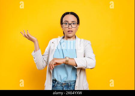 Confuso perplesso ispanico o brasiliana giovane donna, dipendente aziendale, utilizzando smart watch sulla sua mano, preoccupato per essere in ritardo, si trova su sfondo isolato arancione, guarda la delusione fotocamera Foto Stock