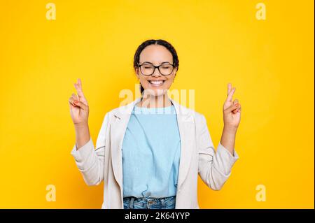 Eccitato positivo brasiliano o latino giovane donna, elegantemente vestita, tenendo le dita incrociate, gli occhi chiusi, facendo desiderio, in attesa di qualcosa di speciale, in piedi su sfondo isolato arancione Foto Stock