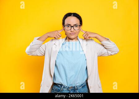 Frustrata turbata brasiliana o ispanica giovane donna brunetta con occhiali, operaio aziendale, coprire le orecchie con le dita, tristemente guarda la macchina fotografica, in piedi su sfondo isolato arancione Foto Stock