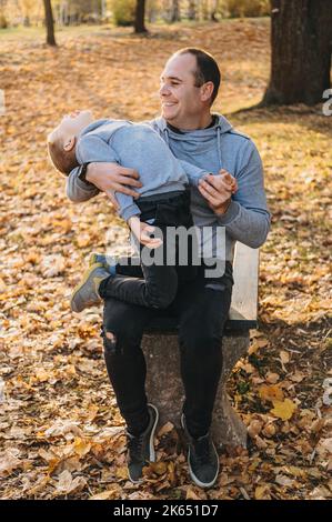 Felice padre seduto sulla panca di legno nel parco tenendo in braccio il figlio inquieto. Concetto di famiglia, infanzia, stagione e persone. Foto Stock