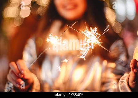 Giovane donna che tiene gli scintillatori mentre si trova in piedi contro le luci di Natale in città Foto Stock