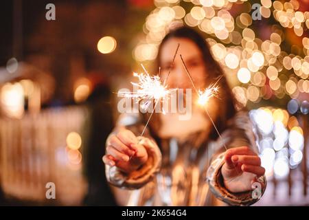 Giovane donna felice che tiene gli scintillatori mentre si trova contro l'albero di Natale illuminato all'aperto in città Foto Stock