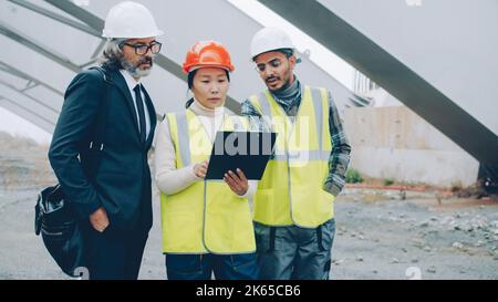 Il gruppo multietnico di architetti sta leggendo documenti e progetti che parlano in piedi in cantiere insieme discutendo il progetto di costruzione. Foto Stock