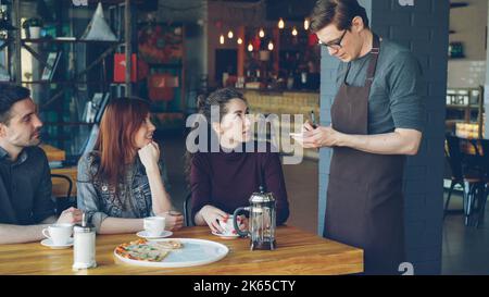 Il cameriere maschile amichevole sta prendendo ordine da un allegro gruppo di amici seduti al tavolo in un moderno caffè e parlando. Mangiare fuori, industria alimentare e delle bevande e concetto di amicizia. Foto Stock