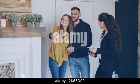 Agente immobiliare è incontro felice coppia sposata, mostrando documenti e raccontando loro di nuovo appartamento. I giovani sono eccitati, guardano intorno e abbracciano. Foto Stock