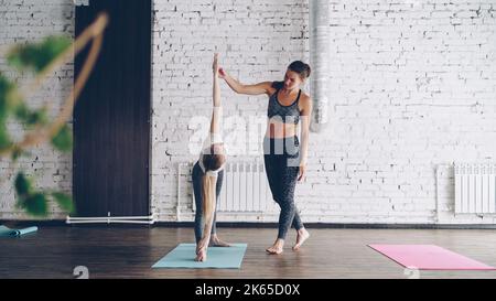 Insegnante di yoga femminile con esperienza sta dimostrando una posa triangolare estesa a una giovane studentessa bionda e correggendo la posizione del suo corpo e del suo braccio durante la pratica individuale nel centro benessere. . Foto Stock