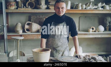 Ritratto di bel giovane vasaio in grembiule fangoso in piedi a tavola in officina vasaio e guardando la macchina fotografica. Ripiani con vasi e pentole fatti a mano sullo sfondo. Foto Stock