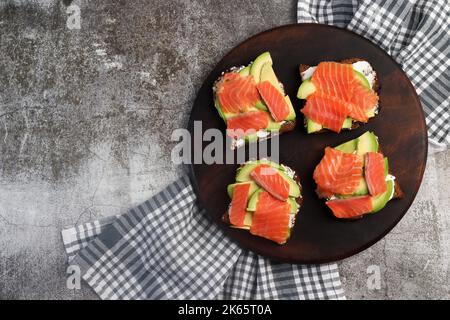 Panini all'avocado al salmone fatti in casa con formaggio casolare su un tagliere di legno rotondo su sfondo grigio scuro. Vista dall'alto, disposizione piatta Foto Stock