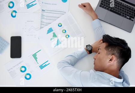 Primo piano di un giovane uomo d'affari asiatico stanco che dorme su un tavolo pieno di documenti e tecnologia wireless. Giovane uomo che guarda esausto con un notebook Foto Stock