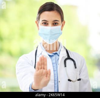 Ritratto di una donna ispanica medico che indossa una maschera e mostra un gesto di arresto con la mano mentre indossa uno stetoscopio in un ufficio ospedaliero. Foto Stock