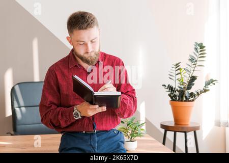 Pianificazione del budget. Uomo pensive barbuto che scrive in un notebook mentre seduto al tavolo in un ufficio moderno in abiti di lavoro, business planning concetto Foto Stock