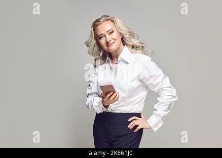 Elegante donna di mezza età con capelli biondi che tiene il telefono in piedi su un grigio isolato Foto Stock