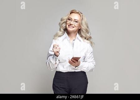 Donna d'affari di mezza età con occhiali rigidi, tiene lo smartphone su uno sfondo grigio. Foto Stock