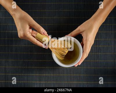 Le mani preparano il tè di Matcha in una ciotola bianca con una frusta di bambù Foto Stock