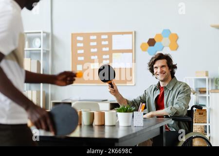 Ritratto di giovane uomo sorridente con disabilità giocando a ping pong in ufficio sala pausa contro il collega, copia spazio Foto Stock