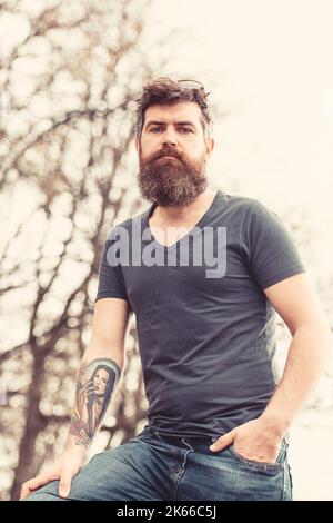 Uomo con barba e baffi sul volto stretto, alberi sullo sfondo, sfocato. Uomo bearded in abito casual in posa all'aperto. Uomo con barba lunga Foto Stock