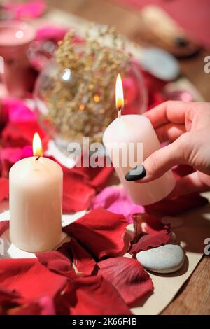 mano femminile che tiene la candela bruciante sopra i petali di rosa, le pietre e le erbe stratificate su un tavolo. letture di aura e rituali magici, spiritualistic e channelle Foto Stock