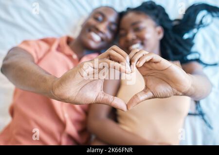 Coppia afro-americana che mostra un gesto a forma di cuore con le mani e sorridente in uno sfondo sfocato mentre sdraiato su un letto a casa. Giovane uomo nero Foto Stock