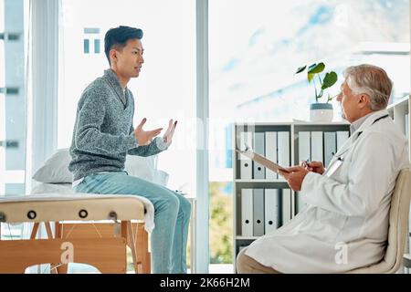 Un giovane asiatico che parla mentre si consulta con un medico caucasico senior mentre si siede su un letto in clinica. Paziente di razza mista che spiega come lui Foto Stock