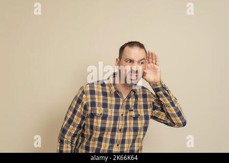 L'uomo che ascolta tiene la mano vicino all'orecchio su uno sfondo beige. Bell'uomo che cerca di ascoltare la conversazione di qualcuno Foto Stock