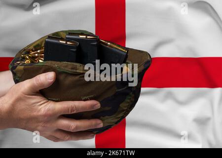 Un casco militare con cartucce e riviste per un fucile nelle mani di un uomo sullo sfondo della bandiera inglese. Il concetto di Foto Stock