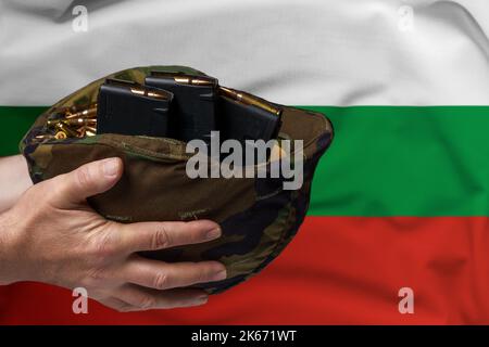 Un casco militare con cartucce e riviste per un fucile nelle mani di un uomo sullo sfondo della bandiera di Bulgaria. Il concetto di Foto Stock