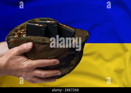 Un casco militare con cartucce e riviste per un fucile nelle mani di un uomo sullo sfondo della bandiera Ucraina. Il concetto di Foto Stock