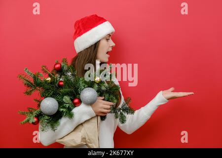 Stupito scioccato giovane donna che tiene bouquet di rami di abete rosso che presenta con palma area di lavoro copia spazio, indossando maglia bianca maglione Santa cappello, Foto Stock