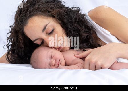 la madre abbraccia e guarda teneramente il suo neonato mentre dorme e si riposa sul letto nella stanza. Sfondo bianco. Concetto di famiglia e di affetto Foto Stock