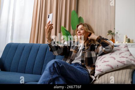 Donna matura bionda che prende selfie sullo smartphone mentre si siede sul divano in appartamento Foto Stock