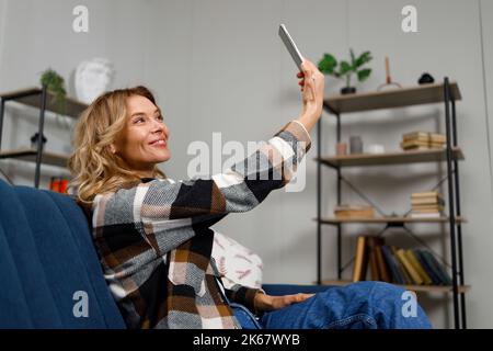 Donna sorridente di mezza età che porta selfie sullo smartphone mentre si siede sul divano di casa Foto Stock