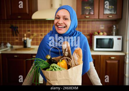 Bella donna musulmana sorridente casalinga in hijab in piedi con una borsa di spesa di alimentari contro una cucina di legno a casa Foto Stock