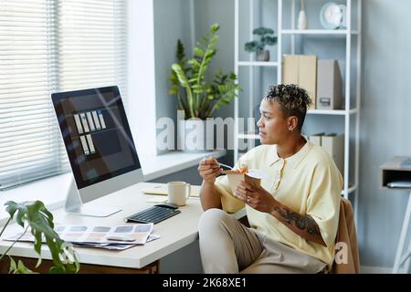 Vista laterale ritratto di una donna nera tatuata che mangia cibo da asporto sul posto di lavoro durante la pausa pranzo e guardando lo schermo del computer Foto Stock