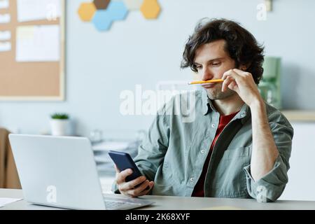 Ritratto del giovane caucasico che gioca con la matita e usa il telefono sul posto di lavoro che soffre di noia e procrastinazione Foto Stock