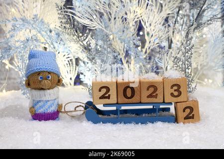 Un gnomo giocattolo lavorato a maglia si trova sulla neve artificiale e una slitta giocattolo con cubi di legno con i numeri 2023 si trova accanto a lui sullo sfondo di Foto Stock