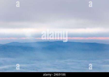 Raggi di sole che scende da alcune nuvole sopra una valle piena di nebbia, illuminando una parte di esso Foto Stock