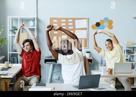 Gruppo eterogeneo di giovani sorridenti che fanno esercizi stretching sul posto di lavoro in ufficio Foto Stock