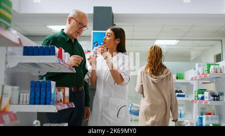 Farmacista asiatico che spiega farmaci cardiologici all'uomo anziano, parlando di medicina sanitaria in farmacia. Donna che aiuta il cliente con trattamento e farmaci, assistenza medica. Foto Stock