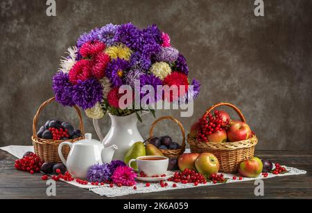 Autunno ancora vita. Un vaso con assaggiatori, cesti di frutta, mele e prugne e porcellana in teaware sul tavolo Foto Stock
