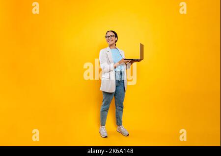 Foto a tutta lunghezza di felice successo elegante latino o brasiliana giovane donna, dipendente d'affari, con occhiali, tenere in mano il portatile aperto, stare in piedi su sfondo isolato arancione, guarda al lato, sorriso Foto Stock