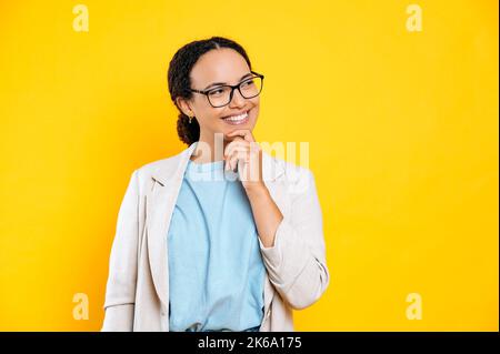 Felice sognante gorgeous latino o brasiliana giovane bruna donna d'affari, con gli occhiali, tiene la mano vicino al mento, guardando da parte, sognando, progettando, sorridendo, si trova su sfondo arancione isolato Foto Stock