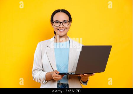 Positivo sicuro intelligente bella latino o brasiliana giovane bruna donna, donna d'affari, con gli occhiali, tenere in mano il portatile aperto, stare in piedi su sfondo arancione isolato, guarda la fotocamera, sorrisi amichevole Foto Stock