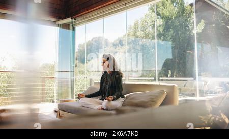 Guaritore olistico sorridendo mentre meditava su un divano nel suo salotto. Donna anziana felice pratica hatha yoga a casa. Donna spirituale che pratica un Foto Stock